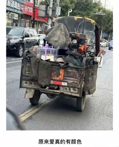 这是一张城市街头的实拍照片，画面主体是一辆外观破旧、沾满污渍的货运三轮车，车厢内杂乱堆放着脏污的工作布料、机械部件等工具，与这些粗糙的物品形成强烈反差的是，车厢里放置着一个包装精美的透明蛋糕礼盒，看起来是为某个重要的人准备的礼物。图片下方配文“原来爱真的有颜色”，通过简陋的劳动工具与精致礼物的对比，传递出即使在平凡朴素的底层生活中，也存在着细腻温暖的爱意，可能是劳动者为家人准备的生日或纪念日惊喜，凸显了爱的纯粹与生活的烟火气。