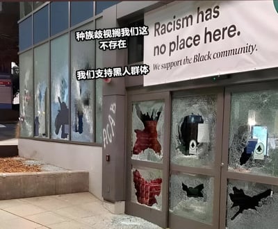 图片展示了一栋建筑的外立面，其玻璃门窗大面积破碎，散落着玻璃碎片。建筑上方悬挂着一条白色横幅，用英文写着"Racism has no place here. We support the Black community."（种族歧视在这里没有立足之地。我们支持黑人群体。）。图片左侧叠加了两行中文文字："种族歧视搁我们这不存在"和"我们支持黑人群体"。破碎的门窗与倡导反种族歧视的标语形成鲜明对比，具有强烈的讽刺意味，暗示了表面口号与实际情况之间的矛盾。