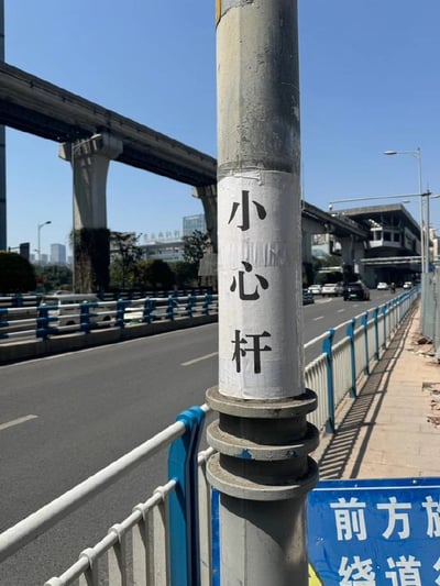 这是一张在城市道路旁拍摄的实拍图，晴朗白天的户外场景中，画面主体是一根立在道路护栏旁的金属杆子，杆子上贴着一张白色贴纸，上面写着“小心杆”三个大字，这是一个谐音梗，谐音“小心肝”，既起到提醒路人注意避让杆子的作用，又制造出趣味效果。背景中可以看到城市高架桥、行驶的车辆和城市建筑，杆子下方还有一块蓝色的提示牌，露出的文字为“前方放 终道”，推测完整内容应为“前方施工 占道”。整体场景充满日常趣味，是网友捕捉到的生活化搞笑细节。