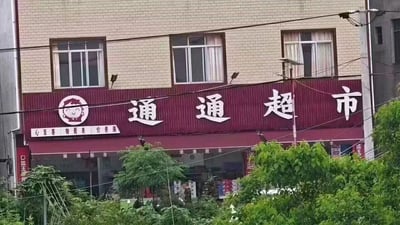 图片展示了一家名为“通通超市”的店铺外观。建筑为两层砖墙结构，招牌为深红色，上方有白色大字“通通超市”，左侧配有一个卡通人物头像logo。招牌下方有一行小字“心至善 物最美 价更廉”。店铺前有绿色植物，背景可见窗户和电线，整体呈现出日常街边超市的真实场景。
