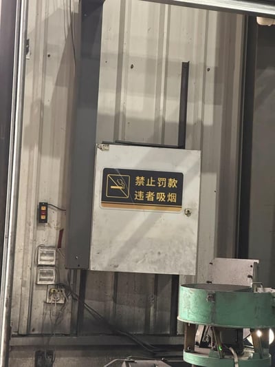 这是一张实拍的工厂车间场景照片，画面中央的白色电气箱体上贴着一块黑底标识牌，标识牌带有标准的禁止吸烟图标，但文字内容顺序完全颠倒，原本规范的标识应该是“禁止吸烟 违者罚款”，此处错误写为“禁止罚款 违者吸烟”，产生了违背常理的搞笑反差效果；画面左侧可见厂房波纹墙面、裸露的电线和电源控制按钮，右下角摆放着一台绿色的工业生产设备，整体是工厂内部的日常场景，出错的标识是画面的核心内容。