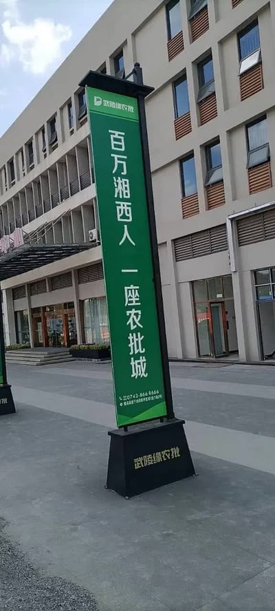 这是一张室外实拍的竖版静态照片，画面主体为一块立在灰色广场地面上的立式宣传导视牌。导视牌底座为黑色，印有金色的“武陵缘农批”字样，牌身主体为绿色，顶部带有武陵缘农批的品牌标识，中间竖排印有醒目的白色宣传大字“百万湘西人 一座农批城”，底部印有项目联系电话及相关地址提示信息。导视牌后方是一栋浅米色的多层商用建筑，楼体分布有整齐排列的窗户，底层为商铺入口，建筑前方的天空能看到少量白云，整体是农批城项目的线下宣传场景。