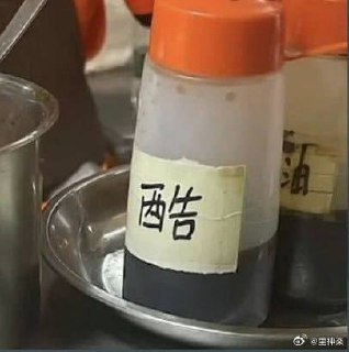 厨房场景中，一个带橙色盖子的透明塑料容器放在金属托盘上，瓶身贴有手写白色标签，上面用黑色马克笔写着‘西告’二字。容器内装有深色液体，疑似酱油。旁边可见其他类似容器，整体环境像是餐馆或家庭厨房的调料区。

图片中的‘西告’可能是‘酱油’的戏谑性误写，将‘酱’字拆分为‘西’和‘告’，形成谐音或拆字梗，带有幽默效果。