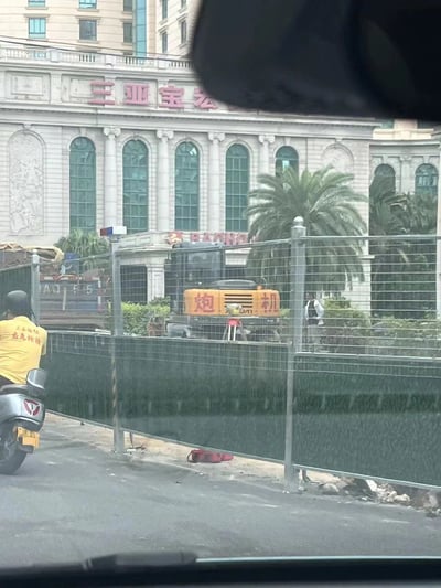 从车辆内部拍摄的三亚街景，背景为带有“三亚宝宏”字样的大型建筑，前方有绿色施工围栏围起的工地，内有黄色“炮机”挖掘机。左侧一名身穿黄色夹克、头戴安全帽的摩托车手正骑行经过，展现了城市中日常的建设场景与交通画面。