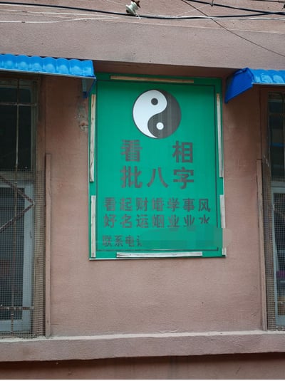 这是一张实拍的老旧居民楼外墙局部照片，浅棕色的水泥墙面上固定着一块绿底灰字的算命服务招牌，招牌顶部印有太极阴阳鱼的道教标识，最上方以较大字号标注核心业务为“看相、批八字”；下方罗列细分业务的文字采用了特殊排版，被排列为横排的两行，乍看横读会得到“看起财婚学事风”“好名运烟业业水”这类不通顺的内容，实际按竖列读取才是正确的业务内容：看好、起名、财运、婚姻、学业、事业、风水；招牌最下方的联系电话部分已做打码模糊处理。招牌两侧是装有蓝色简易雨棚的老旧玻璃窗，墙面上方还可见外露的电线和绝缘子。