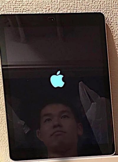一张苹果设备的照片，屏幕上亮着标志性的苹果logo，同时能看到拍摄者的面部倒影。设备放置在一个纹理粗糙的表面上，可能是布料材质。