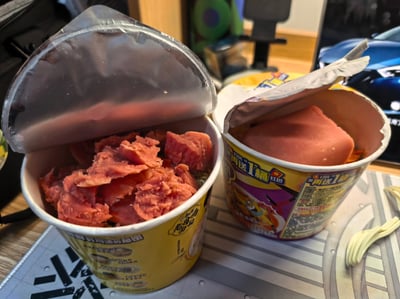 两桶开盖的方便面放在桌面上，左侧桶内堆满撕碎的粉红色火腿，右侧桶内则放着一整块厚实的火腿，桶身包装印有“再送1桶”“红包”等促销字样。背景中隐约可见电脑屏幕上的汽车图片，整体场景像是在展示“豪华升级版”方便面，配文“不准羡慕”充满炫耀意味，让人会心一笑。