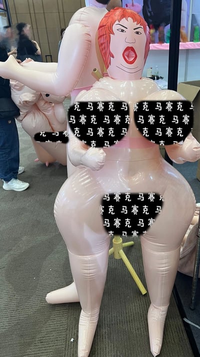 图片展示了一个在展会现场的大型充气人偶，具有夸张的卡通化外形：红色短发、张大的嘴巴和红色嘴唇，以及被多处黑色“马赛克”文字遮挡的身体部位。人偶呈现站立姿势，双臂向两侧张开。背景中可见模糊的展会环境和几位参观者，显示这是在某个展览或活动现场拍摄的照片。