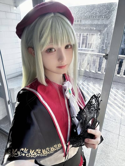 一位银发白皮肤的cosplayer，头戴红色贝雷帽，身穿红黑相间带有金色花纹的服装，手持黑色蕾丝物品，站在玻璃门前的户外场景。背景可见建筑物和阳台栏杆，光线明亮，整体呈现出精致的动漫角色扮演风格。