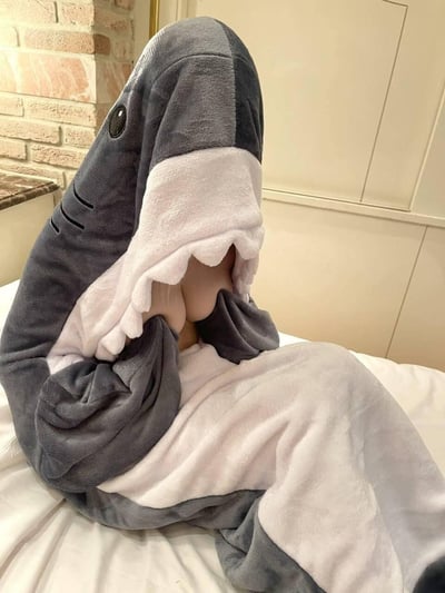 一张生活实拍图，画面里的小朋友裹着灰白带锯齿状“嘴巴”的鲨鱼造型连帽睡袋，把脸藏在鲨鱼的“嘴巴”内部，只露出部分脸颊，坐在铺着白色床单的床上，睡袋看起来毛绒绒的，整体氛围软萌治愈，没有双关或谐音梗内容。