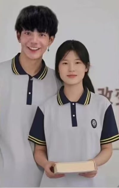 这是一张AI生成的竖版图片，画面中一男一女身着同款浅灰色拼藏青色的Polo领校服，校服领口和袖口带有黄黑条纹装饰，女生左胸口还有校徽样式的标识。男生站在女生身后，露出灿烂的笑容，耳朵上戴着耳饰；女生站在前方，双手捧着一本厚书，面带温和的微笑，头发扎成马尾。背景简洁，右侧能看到模糊的“改”字，整体氛围清新温馨，呈现出校园情侣合照的感觉。
