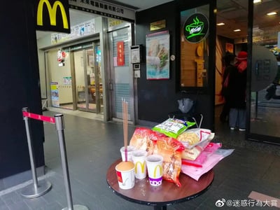 这张图片拍摄于麦当劳门店的入口区域，在门店地面的小桌上，摆放着一套中式祭祀供品：三根长香插在麦当劳的纸杯中，旁边堆叠着多袋零食、点心类食品。画面左侧是麦当劳的排队隔离栏，背景里可以看到麦当劳门店的McCafe区域、旁边的其他商铺，还有贴在门上的“春臨有福”春联，门店内还有顾客在活动。原本是西式快餐门店的场景，搭配中式祭祀供品，形成了反差感强烈的迷惑画面，看起来像是在对麦当劳门店进行中式祭祀。