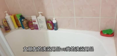 这是一张浴室场景的静态实拍照片，聚焦于浴缸边缘的洗浴用品对比。画面左侧整齐摆放着十多瓶不同品牌、包装样式各异的洗浴产品，涵盖了洗发水、沐浴露、护发素等多种品类，这部分代表女朋友的洗浴用品；而画面右侧的角落中，仅放置着一个红色皂盒，里面只有一块绿色的肥皂，这部分代表“我”的洗浴用品。图片下方配有文字“女朋友的洗浴用品vs我的洗浴用品”，通过两者在数量和品类上的强烈反差对比，生动调侃了情侣间在洗浴用品使用上的巨大差异，内容贴近日常生活，容易引发同类人群的共鸣。