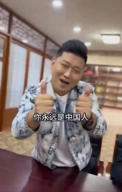 在带有中式木质窗格的室内场景中，一名穿着花纹外套的男子眯起双眼、咧嘴大笑，双手同时竖起大拇指，画面下方配有醒目的字幕“你永远是中国人”，整体氛围热情又接地气，传递出直白的正能量情感，没有双关、谐音梗或隐含暗示内容。