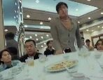 图片展示了一个餐厅场景，一位身着灰色西装的女子正俯身靠近餐桌，桌上摆满了菜肴，周围坐着几位正在用餐或交谈的食客。整体氛围像是在进行一场聚餐或商务宴请。