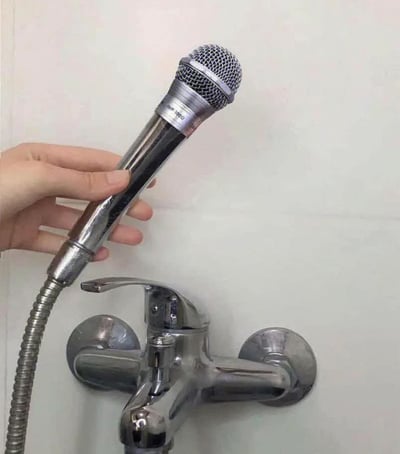 整张图为浴室实拍场景，墙面上安装着银色的冷热水混水阀，连接混水阀的金属波纹花洒软管没有接常规的花洒头，反而安装了一支外观为银色金属质感的动圈麦克风，二者外形和质感高度相似，一只手正握住这支接在花洒管上的麦克风，形成了极具反差的搞笑错位效果，呼应了很多人有洗澡时唱歌的日常习惯，趣味十足。