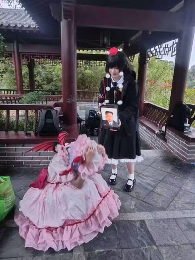这张图片拍摄于户外中式凉亭中，画面里有两位Cosplay爱好者：一位身着粉色多层裙摆、带有红色小翅膀的cos服（还原东方Project中的蕾米莉亚·斯卡雷特），跪在石板地面上，身体前倾做出跪拜的动作，头部被裙摆遮挡无法看清表情；另一位身着黑白色系cos服（还原东方Project中的射命丸文），站在一旁，双手举着一台平板电脑，平板屏幕上显示着一张男性人像。整个场景是coser的整活创作，将二次元cosplay场景与三次元人像结合，通过跪拜的动作制造出荒诞搞笑的反差效果，模仿信徒跪拜的仪式感，属于二次元爱好者的离谱玩梗行为。
