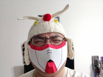 图中人物戴着手工钩织的高达造型针织护耳帽，帽子还原了RX-78-2高达头部的白色天线、红色顶珠和黄色眼部细节，搭配3D打印后上色的高达下半脸面罩，人物皱起眉头眯着眼睛，表情搞怪有趣，是高达爱好者的自制创意cos作品。