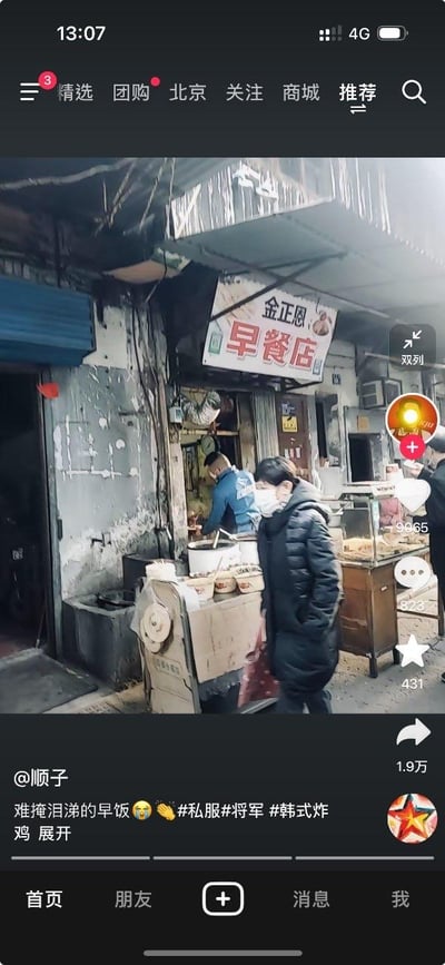 这是一张抖音平台的竖屏短视频截图，画面展示了一家外观老旧的街边早餐店，店铺招牌被恶搞为“金正恩 早餐店”，充满戏谑感。画面中，一位穿黑色长款羽绒服、戴口罩的行人正从店前走过，店内有穿蓝色外套的人在忙碌，餐食操作台摆放着简单的容器，环境充满市井烟火气。视频发布者为@顺子，配文“难掩泪涕的早饭”并搭配表情和#私服#将军#韩式炸鸡 等标签，视频右侧显示互动数据：点赞9065、评论323、收藏431、转发1.9万，界面顶部是抖音的导航栏，底部是抖音的功能栏。