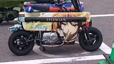 这是一辆本田（HONDA）小型摩托车，被ACG爱好者改装为痛车：车身侧面印有动画《吹响！上低音号》的主角黄前久美子的形象，角色的号嘴位置与摩托车排气管巧妙重合，形成了角色仿佛在吹奏排气管的趣味视觉效果。车身上还印有英文“HONDA”“Kanoniko”，以及日文原名“響け！ユーフォニアム”的字样，车头上方还放置了一个蓝白配色的小玩偶。车辆停放在户外沥青路面上，背景可见其他车辆与道路设施。
视觉巧思：角色号嘴与排气管的位置重合，制造出有趣的画面错位感。