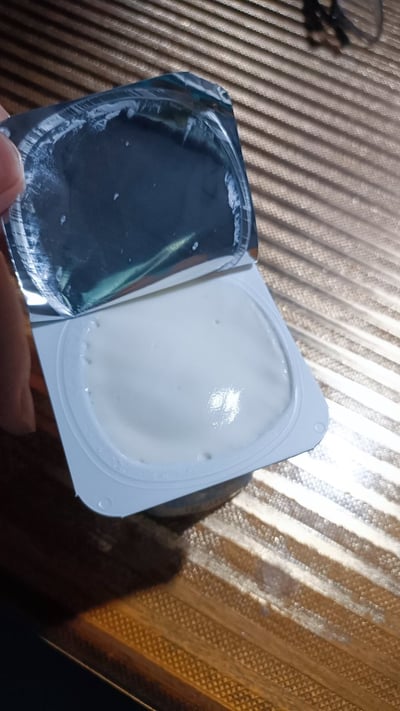 一只手正拿着一个打开的白色塑料容器，容器内装有白色乳状物质，可能是酸奶或布丁。容器的铝箔盖子已被部分揭开，露出内部光滑的表面，盖子上还残留少量内容物。背景是带有平行条纹的金属质感桌面，光线从右上方照射，在容器和桌面上形成明暗对比。