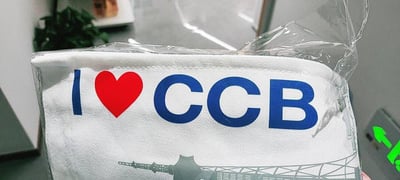 图片近距离展示了一个白色口罩，口罩外覆透明塑料包装。口罩正面印有蓝色的'I'字母、红色爱心图案和蓝色的'CCB'字样，下方还有灰色的建筑轮廓图案。背景中可见模糊的绿色植物和灰色桌面，整体呈现出商品特写的场景。