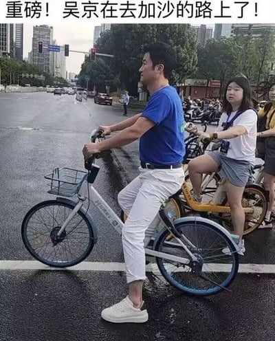 这是一张城市路口的真实拍摄照片，画面里一名穿蓝色短袖、白色长裤的男子骑着哈啰共享单车，他的外形与演员吴京高度相似，在他身后还有一名女子也骑着共享单车。照片上方被添加了恶搞文字“重磅！吴京在去加沙的路上了！”，利用路人撞脸明星的巧合，配上夸张的文字制造出搞笑戏谑的效果，属于典型的网友玩梗内容，没有双关、谐音等其他隐含含义。