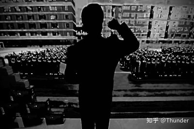 这是一张利用视觉错位原理创作的黑白合成图，前景的小型人物剪影模型站立在高处，抬手做出演讲的姿态，其前方摆放着一些小型道具；背景的居民楼窗户与楼前区域在特殊视角下，恰好形成了整齐列队的“听众人群”与开阔场地的视觉错觉，营造出一种演讲者在宏大场合发言的震撼场景。黑白色调进一步强化了画面的氛围感，让错位带来的错觉更具真实感，巧妙地将日常的居民楼场景转化为充满仪式感的虚构演讲现场。
