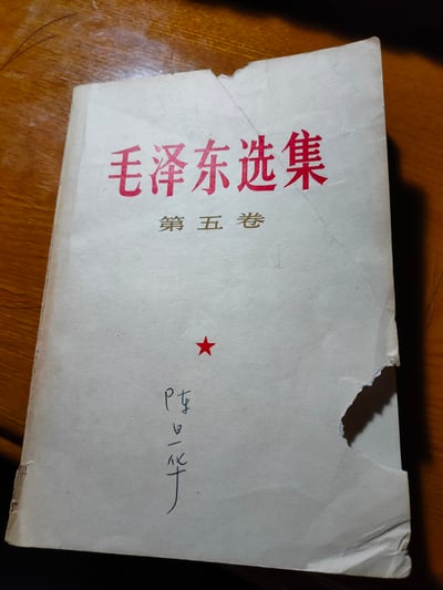 这是一张展示《毛泽东选集第五卷》旧书封面的照片。封面为白色纸张，上方有红色的"毛泽东选集"书名，下方有金色的"第五卷"字样，中间偏下位置有一颗红色五角星。封面右下角有手写的"陈日化"名字。书本边缘有明显磨损、撕裂和折痕，显示出一定的年代感和使用痕迹。