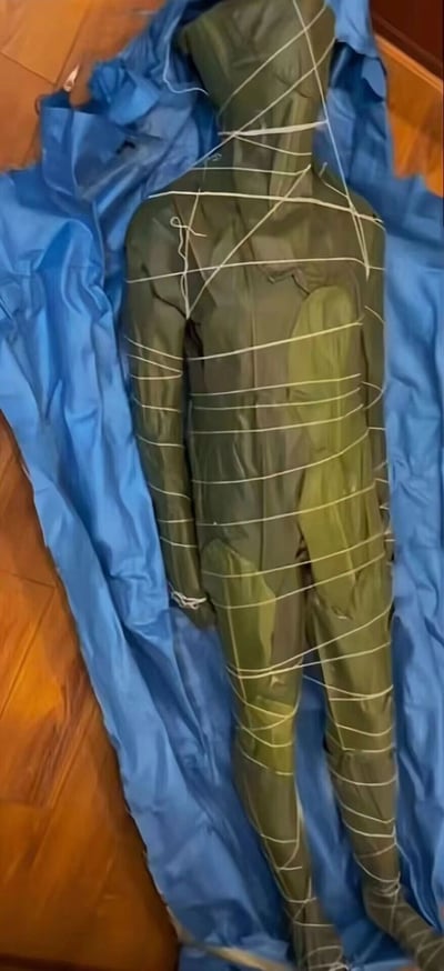这是一张竖屏静态单图，画面主体是一个深绿色的充气人形道具，该道具被白色的绳子以横向、交叉的方式多圈捆绑固定，道具被放置在一块蓝色的布料上，背景是浅棕色的木质地板。这个道具外观模拟人体形态，看起来可以用于户外拓展训练、应急演练中的假人模拟，也可能是作为整蛊类的充气玩具。