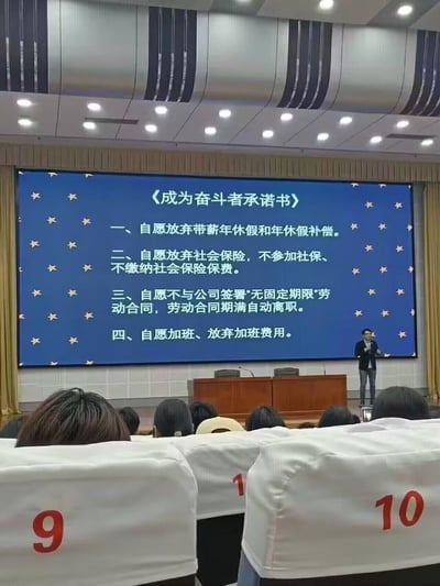 这是一张会议现场的实拍照片，画面中大屏幕展示了一份《成为奋斗者承诺书》，条款内容包括自愿放弃带薪年假及补偿、自愿放弃社会保险、自愿不签署无固定期限劳动合同到期自动离职、自愿加班放弃加班费，这些条款严重违反劳动法律法规，侵害劳动者合法权益。现场台下坐有众多听众，前方有一位宣讲人正在进行讲解，整体场景充满荒诞感，凸显了企业不合理的用工要求。