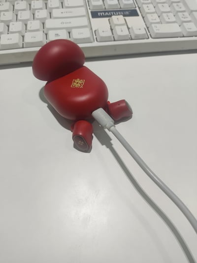 这是一张办公桌面的实拍静态图，画面主体是一个设计成卡通玩偶造型的红色真无线（TWS）耳机充电仓，充电仓正面印有金色中式卷草纹样式的标识，底部插着印有“SIKENAI”字样的白色充电线，处于充电状态。充电仓的“手臂”位置露出收纳的红色耳机，耳机外侧带有金色四叶草形标识。背景是一台白色的MAITU脉途键盘，放置在干净的白色桌面上，整体风格清新可爱，展现日常办公场景里的数码小物。