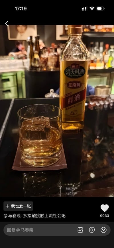 这是一张社交媒体内容截图，拍摄场景为酒吧吧台，黑色台面上的棕色杯垫上摆着一只装有冰块和棕黄色液体的玻璃杯，杯子旁立着一瓶本作为烹饪调味品的海天江南黄料酒，背景可见酒吧酒架、各类酒品以及模糊的店内人员。截图底部配有用户@马春晓的评论“多接触接触上流社会吧”，内容将日常调料料酒伪装成酒吧售卖的高档洋酒，通过强烈的反差制造搞笑效果，反讽了部分人推崇的所谓“上流社会”的虚伪与矫情。