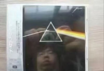 这是一张趣味整活的二次创作图，将英国摇滚乐队平克·弗洛伊德的经典专辑《月之暗面》的棱镜分光封面与真人自拍结合，通过错位拍摄让人物的脸刚好处于封面的棱镜图案后方，同时用道具模拟封面里的光线穿过棱镜的效果，制造出专辑封面与真人融合的搞笑视觉错位，充满搞怪趣味。