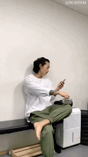 这是一段动态GIF图，画面中一位身着白色上衣、绿色长裤的男子坐在长椅上，一只脚抬起，另一条腿上放着餐盒，他正低头看着手机，同时伸手从餐盒里取食物。由于视觉错位，抬起的脚与下方的木质结构形成了类似“假脚”的错觉，让画面看起来仿佛男子的一只脚是假的并放在腿上，充满了搞笑的视觉反差感，十分有趣。