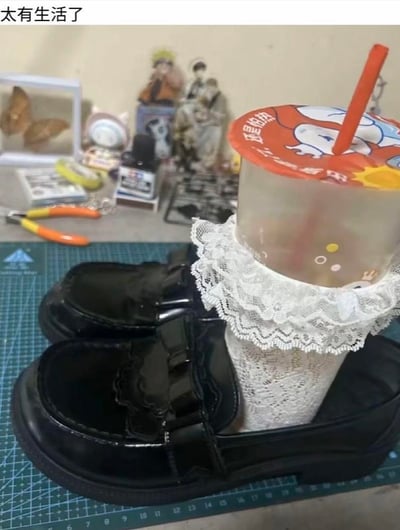 这是一张充满生活创意的视觉错位图，将套着白色蕾丝中筒袜的奶茶杯，放置在一双黑色亮面乐福鞋的鞋腔中，从视觉上营造出“奶茶杯变成脚穿进鞋子”的趣味错觉。画面顶部配有文字“太有生活了”，奶茶杯盖上印有“还是怕热”的字样，整体用日常物品组合出搞笑又有脑洞的效果，让人忍俊不禁。