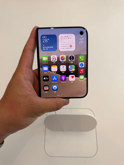 这是一张竖幅原始实拍照片。画面左侧露出一只成年人的左手，手腕处有编织表带，手掌托住一台黑色圆角折叠屏手机，拇指位于底部，手机正面朝向镜头。屏幕处于点亮状态，显示与 iOS 近似的主屏幕：左上角是蓝色天气小组件，可见“中坜 28°”以及强烈季候风信号等内容；旁边是日历小组件，显示 10 月、九月十四和日期表。屏幕中可见 FaceTime、日历、照片、相机、邮件、备忘录、时钟、地图、TV、App Store、Podcast、健康、钱包、搜索等图标，右侧竖向排列电话、Safari、音乐、信息四个 Dock 图标，底部有页面指示点。屏幕右上角有一个单挖孔前置摄像头，时间显示为 12:03。壁纸为沙漠山脉风格，照片中可以看到屏幕折痕造成的反光。背景是干净的浅色墙面和桌面，手机下方有一个白色圆角的陈列或充电类物件。原图文案称折痕实际看并不明显，但用照片拍摄后会显得更清楚。
