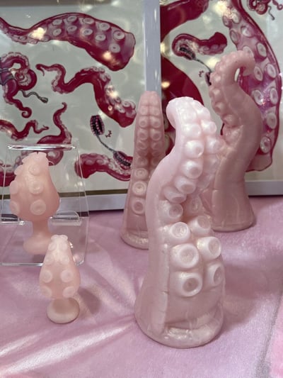 图片展示了一组粉色章鱼触手造型的摆件，有大有小，放置在粉色缎面背景上。较大的触手雕塑呈现出弯曲的形态，表面有逼真的吸盘纹理；较小的摆件则被放置在透明的展示盒中。背景中可以看到印有红色章鱼触手图案的海报，与前景的粉色摆件形成色彩对比。整体场景像是在展示某种手工艺品或特色商品。