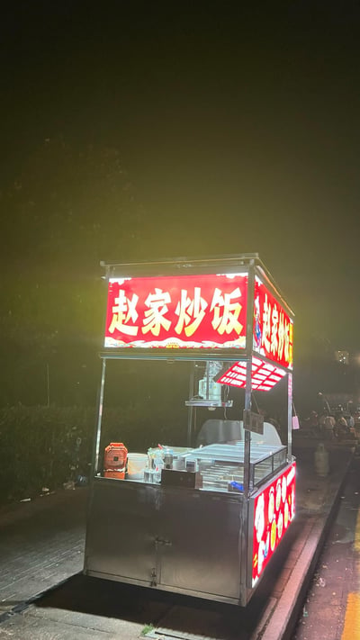 深夜的街道旁，一个亮着暖黄色灯光的炒饭小摊格外显眼。红色招牌上用黄色大字写着“赵家炒饭”，金属制的摊位结构在灯光下泛着冷光，内部摆放着炒锅、食材容器和一台橙色小风扇。周围环境昏暗，隐约能看到远处有行人，营造出城市夜生活中烟火气十足的氛围。