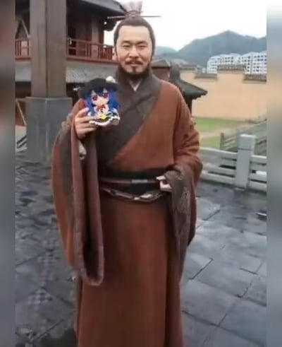 图片中一名身着传统汉服的男子站在古建筑庭院中，背景可见传统木质结构建筑和远山。男子面带微笑，右手持有一个现代动漫风格的蓝色头发少女手办，形成鲜明的古今对比。