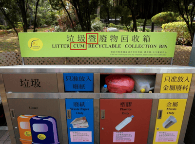 图片展示了一个位于公园环境中的多格垃圾暨废物回收箱。回收箱顶部的绿色标牌上有中文“垃圾暨廢物回收箱”及英文翻译“LITTER CUM RECYCLABLE COLLECTION BIN”。其中“暨”字被错误翻译为“CUM”，而“cum”在英文中具有不雅含义，正确翻译应为“AND”或“WITH”。回收箱分为垃圾、废纸、塑料、金属等投放口，箱体为银色金属材质，背景有树木和绿植，环境整洁。