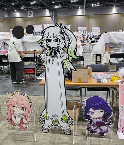 这是一张在同人漫展现场拍摄的照片，画面核心是三个《原神》的同人立牌：中间是尺寸夸张的魔改大慈树王造型纳西妲立牌，角色双手叉腰，表情严肃又带着滑稽感；左右两侧是Q版的八重神子和雷电将军立牌，二者都做出wink的俏皮表情，单手轻靠脸侧。背景是漫展的摊位区域，可见摆放的桌椅、待整理的周边商品和工作人员，整体呈现出同人展会的热闹氛围。