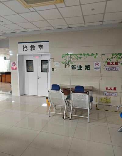 这是一张医院内部的实拍照片，画面左侧是带有“抢救室”黑色标识的白色双开医疗门，门边贴有“抢救重地 闲人免入”以及禁止烟火的警示标识。抢救室右侧的浅色瓷砖墙面上绘制有绿色藤蔓装饰，下方标注着绿色的“作业吧”字样，墙面还配有禁止吸烟标识、消火栓和灭火器的说明提示。作业吧前方摆放着两把带有蓝色靠垫的白色网状输液椅，椅子侧边配有输液架，地面为光洁的浅色瓷砖，整体为医院的公共就诊区域。该作业吧应该是供输液的学生患者在就诊期间书写作业使用，抢救室和作业吧相邻的布局形成了强烈的反差，被网友用来调侃当下学生学业压力过大，即便生病就医也无法耽误写作业的现状，甚至衍生出“写作业快把人送进抢救室”的讽刺性吐槽。