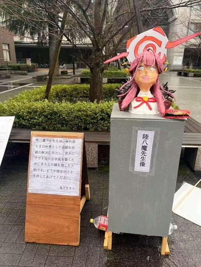 这是在日本京都大学校园内的户外场景，有人手工制作了手游《蔚蓝档案》中角色陆八魔亚瑠的搞怪半身雕像，雕像吐舌的表情十分滑稽，放置在标注有“陆八魔先生像”的灰色台座上。旁边的木质说明牌用日文写着玩梗的介绍，称陆八魔亚瑠作为便利屋68社长为京大解决委托、建立学风，恳请大家原谅她，落款是同游戏角色鬼方佳代子。现场地面潮湿，周围有绿植和树木，是典型的校园户外区域，属于学生的趣味整活行为。