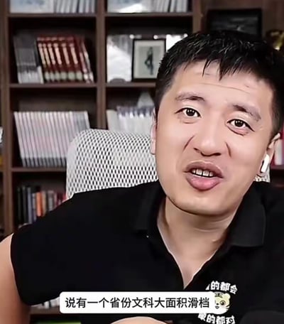 这是一张张雪峰的短视频/直播截图，画面中张雪峰坐在白色网布靠背椅上，身后是摆满各类书籍的深色书架，他佩戴白色蓝牙耳机，身穿带有卡通老虎图案和文字的黑色Polo衫，面部露出惊讶疑惑的神态，似乎正在就高考相关话题发表观点，图片下方配有白色底的字幕，内容为“说有一个省份文科大面积滑档”。