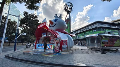 照片展示了一个巨大的蜜雪冰城雪人雕像门店，位于城市街道旁。雪人雕像戴着皇冠，围着红色围巾，面带微笑，右手高举饮料。雕像底部是红色屋顶的售卖亭，上面有“蜜雪冰城”的招牌和招聘信息。背景中可以看到现代建筑、树木和蓝天白云，一名顾客正在亭前选购饮品。整个场景色彩鲜艳，充满夏日气息，巨大的雪人雕像成为街头的吸睛地标。