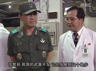 这是一张韩国MBC电视台播出的军事题材影视剧截图，画面中左侧是身着深绿色军装、佩戴军帽与眼镜的军官，右侧是穿着白色医生大褂、挂着工作牌的男性医生，二人身处布满仪器设备的室内环境，看起来像是武器研发相关的场所。画面底部配有中文字幕：“没想到 我国的武器开发 已经发展到这个地步”，传递出人物对本国武器研发进度的惊叹与感慨。