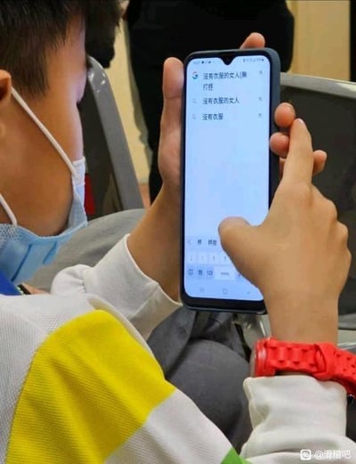 图中是一位戴着蓝色口罩的小孩，身处类似公共候车的场所，正双手操作智能手机。手机屏幕的输入法界面显示，输入框内的内容为“没有衣服的女人(摔打)”，候选词栏呈现“没有衣服的女人”“没有衣服”两个候选选项，画面记录了生活里普通又带有趣味的小场景，不存在双关、谐音梗或暗示内容。