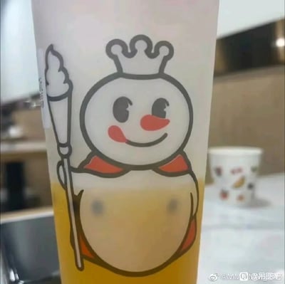 这是一张蜜雪冰城茶饮的实拍图，杯身印有品牌标志性的雪人IP形象，雪人面带微笑、手持冰淇淋权杖，造型可爱。杯子中装有黄色的茶饮，其中的两颗黑色珍珠恰好位于雪人腹部图案的对应位置，形成了类似胸部的视觉错位效果，让原本可爱的雪人形象产生了滑稽搞笑的反差感，属于网友偶然捕捉到的趣味视觉巧合，这类内容常被作为搞笑素材在社交平台分享。