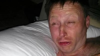 图片中显示一名男子躺在床上，头枕着白色枕头，眼睛红肿，表情悲伤，似乎正在哭泣。背景较暗，主要聚焦于男子的面部表情和上半身。